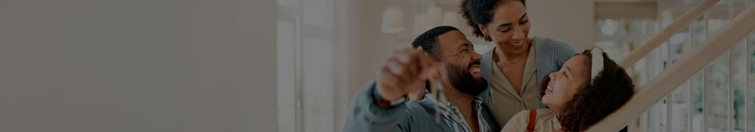 Vista panorâmica de uma cozinha em conceito aberto e sala de estar. Do lado esquerdo a cozinha vários armários, um cooktop, uma pia e uma mesa com três cadeiras, lado direito tem um sofá de dois lugares, travesseiros e dois vasos de plantas.