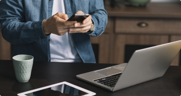 Um homem atras de uma mesa mexendo no celular. Em cima da mesa existem um tablet, um notebook e um copo.